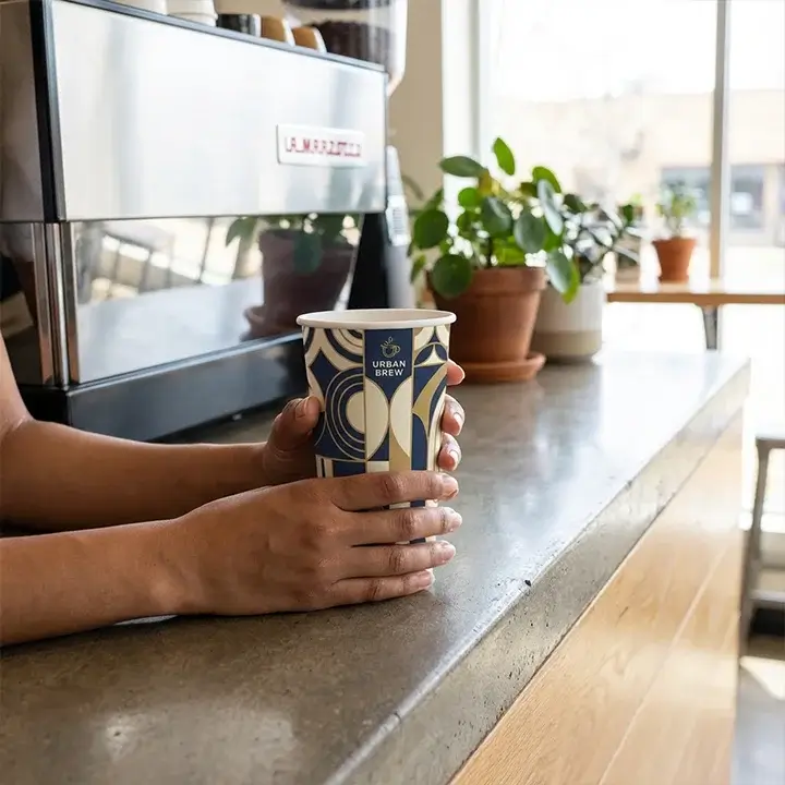 Person holding a custom printed 20 oz paper cup with a branded logo at a modern coffee shop counter